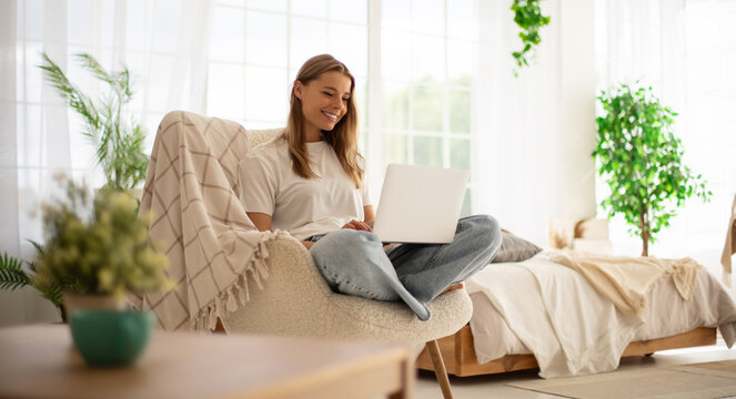 Beautiful young woman sits comfortably in a bright, modern room, working on a laptop. She is seated in a cozy chair with a blanket, surrounded by greenery, natural light, and a relaxed atmosphere.