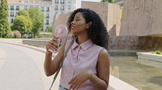 A woman holds a pink handheld fan, cooling herself in a sunny park. She stands near a reflective pond, surrounded by trees and buildings in an urban setting. 