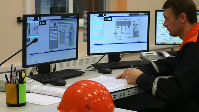 Engineer is managing complex industrial system in control room while multitasking with paperwork and computer screens. Engineer in industrial control room is filling out paperwork while monitoring