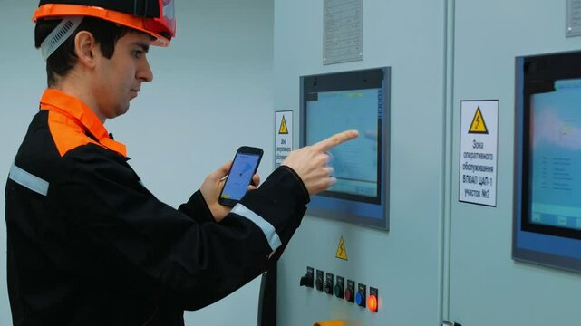 Engineer operates control panel with mobile phone in industrial facility. Engineer wearing hardhat and safety uniform is using mobile phone, touching screen on control panel in industrial facility