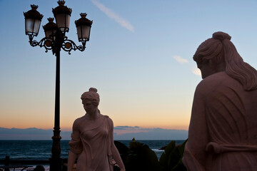 Artistic marble statues facing an ornate street lamp near the ocean, bathed in the warm glow of sunset, creating a romantic and historic ambiance by the seaside.