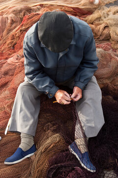 An elderly man in a cap and jacket is sitting on a pile of fishing nets, carefully mending one of the nets with his hands as he focuses on the task at hand in Andalusia Spain