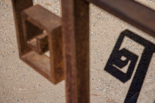An artistic photograph of rusty metallic structures casting geometric shadows on a sandy surface, illustrating the interplay between light, shadow, and texture.