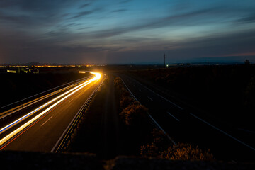 High at night with striking long exposure light trails from vehicle headlights and taillights. The sky is filled with dramatic, dark storm clouds, creating a moody and dynamic atmosphere.