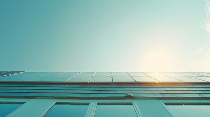 Obraz premium Viewed from below, the bright sun peeks over the sleek, reflective surface of a building covered in solar panels, set against a clear blue sky.
