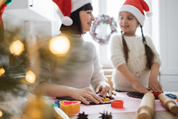 Mother and young daughter in Christmas hats baking cookies together in cozy kitchen. Festive atmosphere with cookie cutters and dough, sharing joyful holiday moments and traditions.