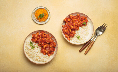Two portions of chicken curry garnished with fresh coriander served with white basmati rice on a yellow background with turmeric and cutlery