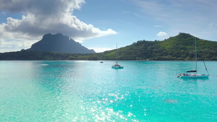 Sailboats anchored in turquoise lagoon, green mountains tropical island in background. Crystal...