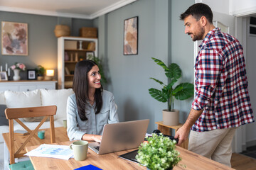 Young business woman working at home from home office on laptop while her boyfriend walking around apartment. Couple goals, successful female freelance worker small company owner do her online job.