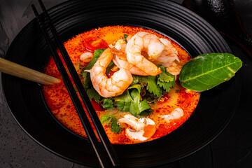 Spicy Thai Tom Yum Kung soup with shrimp, lemongrass and galangal, cilantro, cherry tomatoes and mushrooms in a black bowl on a graphite background with chopsticks and a spoon. Close-up.