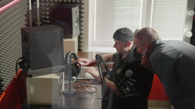 Two men are in a studio room, one seated in front of a computer, likely a producer, wearing black clothes and a cap, while the other, dressed in a long sleeve shirt, leans over, explaining