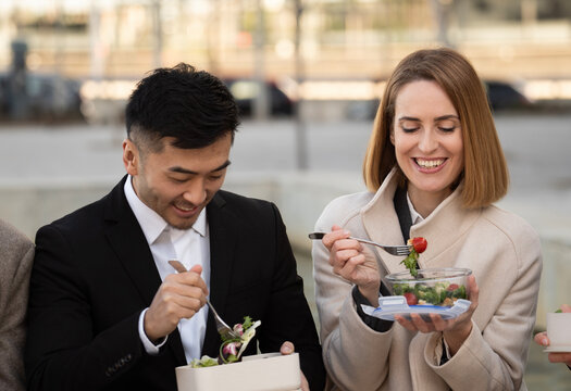 Business man and woman colleagues are eating a healthy salad together on a lunch box outdoors on a lunch break