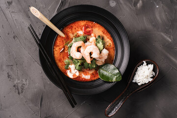 Spicy Thai Tom Yum Kung soup with shrimp, lemongrass and galangal, cilantro, cherry tomatoes and mushrooms in a black bowl on a graphite background with chopsticks and spoon