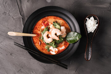 Spicy Thai Tom Yum Kung soup with shrimp, lemongrass and galangal, cilantro, cherry tomatoes and mushrooms in a black bowl on a graphite background with chopsticks and spoon