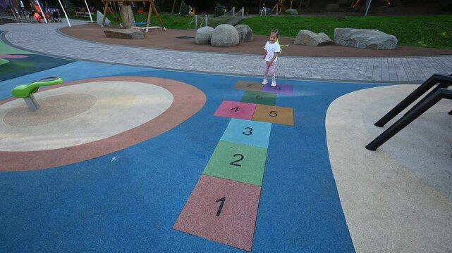 A happy little girl with colorful braids plays hopscotch in a playground. She jumps and smiles at the camera.