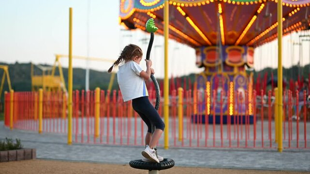 A little girl is playing on a merry-go-round in a playground. She is having fun and laughing. The playground is full of children and their parents.
