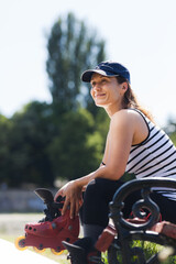 A smiling young woman enjoys a sunny day in the park, sitting on a bench with her inline skates in...