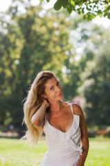 A graceful blonde woman in a stylish white dress poses naturally in a verdant park, with sunlight filtering through trees.