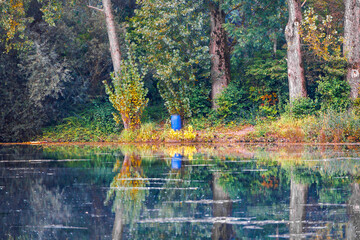 A blue plastic bin for chemicals stands under trees on the bank of a body of water and is reflected in the water