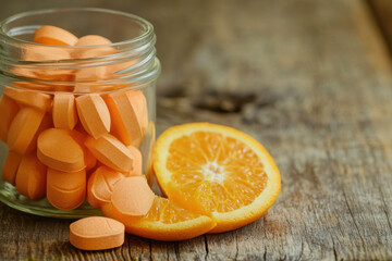 A close-up of vitamin D tablets in a glass jar with a slice of orange beside it, on a rustic wooden table, with blank space on the right for text or an advertisement.