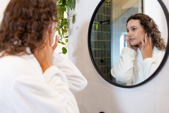 Applying skincare, woman in white robe looking at herself in bathroom mirror