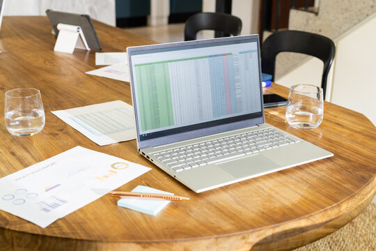 Laptop displaying spreadsheet on wooden table with documents and water glasses