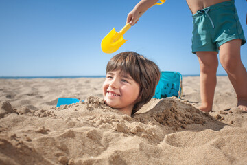 A smiling child's head protrudes from the sand and behind it another unrecognizable child with a...