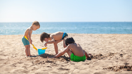 Three children dig a hole in the sand with toy shovels and a bucket full of sea water . © OR MIXphoto