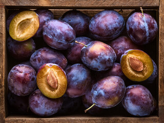 Fresh plums in a wooden crate, close-up of whole and halved purple plums with visible pits.