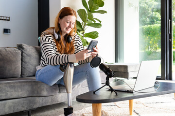 Using smartphone and smiling, woman with prosthetic leg near laptop and microphone