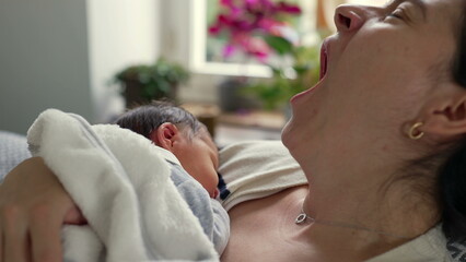 Mother yawning while holding her newborn baby close, illustrating the exhaustion and tender moments...