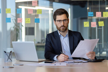 Confident businessman sits at desk reviewing papers with focused expression. Office features glass walls and colorful sticky notes. Scene captures concentration, analysis, and workplace atmosphere.