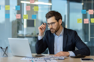Businessman in suit intensely focuses on laptop for data analysis in modern office. Sticky notes on...