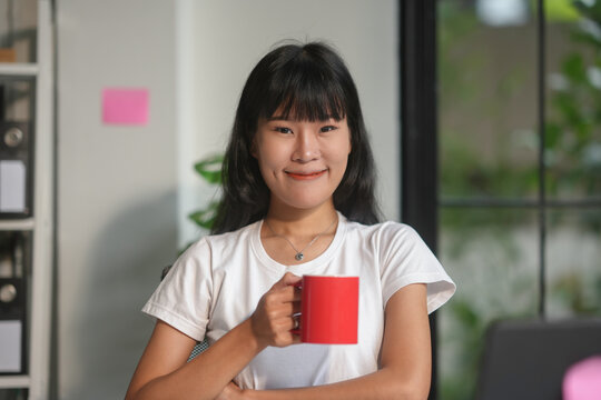 Young asian woman smiles in her office, holding a red mug, surrounded by technology and business essentials, exuding confidence and positivity