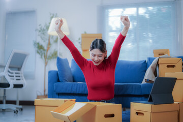 Young woman is sitting on the floor surrounded by cardboard boxes and raising her arms in the air,...