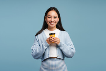 Beautiful woman holding yellow take away coffee cup, looking at camera, standing on blue background