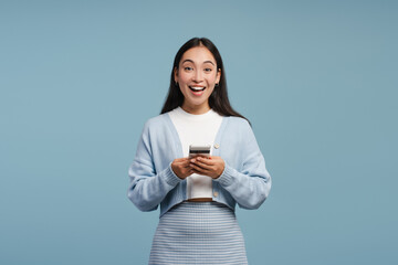 Surprised young Asian woman reading good news on smartphone standing isolated on blue background