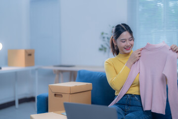 Asian woman sells clothes online from home, showcasing products and engaging with customers through live streaming on her laptop