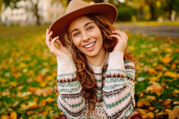 Cheerful young woman wearing a sweater and hat enjoys a sunny autumn day in a park filled with...
