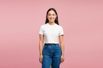 Smiling, attractive Asian woman smiling wearing white t-shirt and jeans on pink background