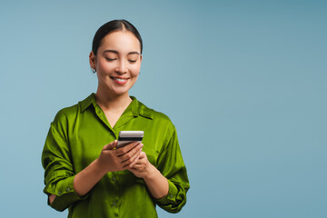 Smiling Asian woman holding mobile phone, using mobile app, standing isolated on blue background