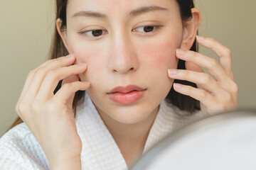 Asian young woman looking face skin in the mirror have a red rash on her face from cosmetic allergy