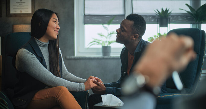 Happy multiethnic couple sit on chairs holding hands and hugging, talk, find compromise after fight on couples therapy. Close up of therapist looking at tender young family on counseling session.