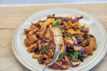 A close-up shot of a fork holding a piece of tofu above a plate of colorful stir-fried vegetables, showcasing a delicious vegan meal.