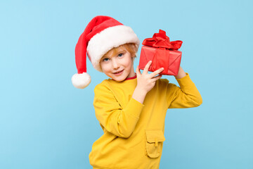 Cute boy wearing red santa hat smiling and holding wrapped christmas present, isolated studio portrait over pastel blue background.