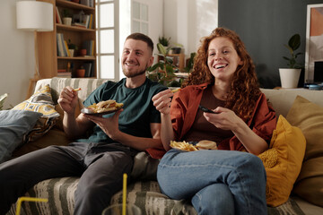 Young cheerful woman with junk food pressing buttons on remote control while choosing channel on tv...