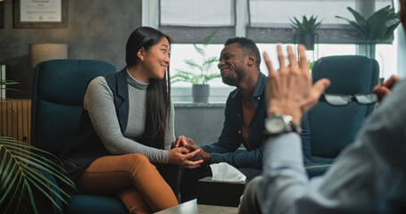 Happy multiethnic couple sit on chairs hand in hand, laugh, look at each other, find compromise...