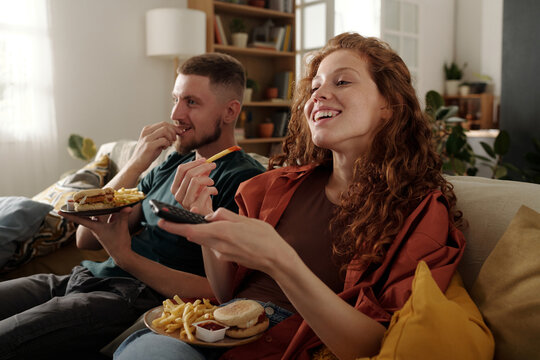 Young smiling woman with french fries eating junk food with her boyfriend and using remote control while watching movie on tv