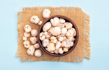 Top View of Makhana or Fox Nut in a Wooden Bowl on Burlap Fabric Isolated on Light Blue Background with Copy Space, Also Known as Lotus Seed Pop, Euryale Ferox or Gorgon Nut