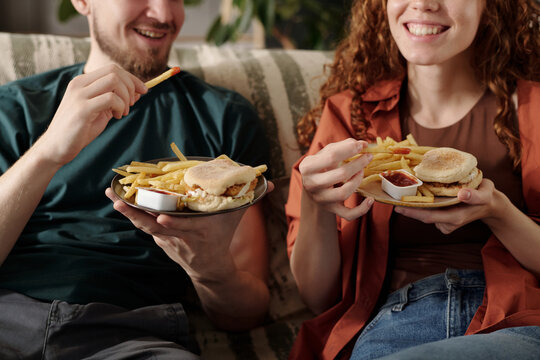 Cropped shot of happy young couple holding plates with french fries and hamburgers while watching tv or home cinema at leisure